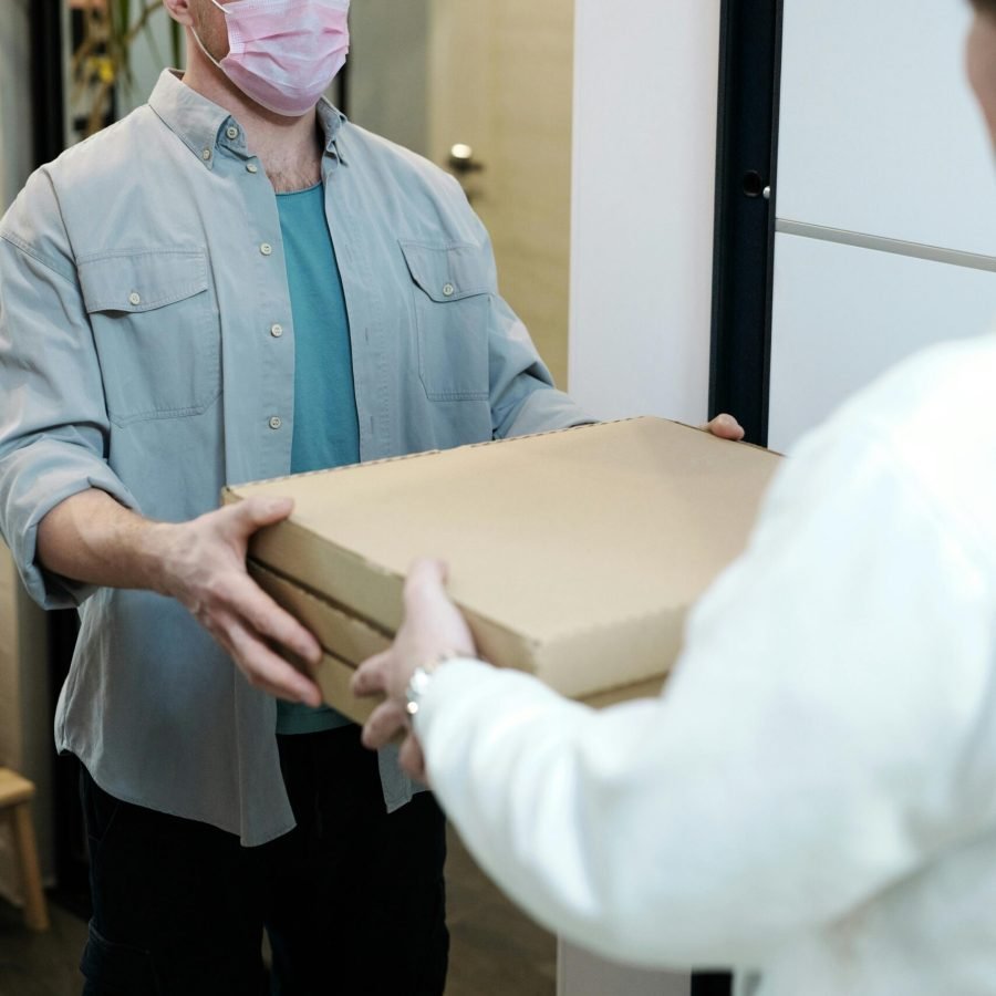 A delivery man with a mask handing over pizza boxes indoors, embodying pandemic safety measures.